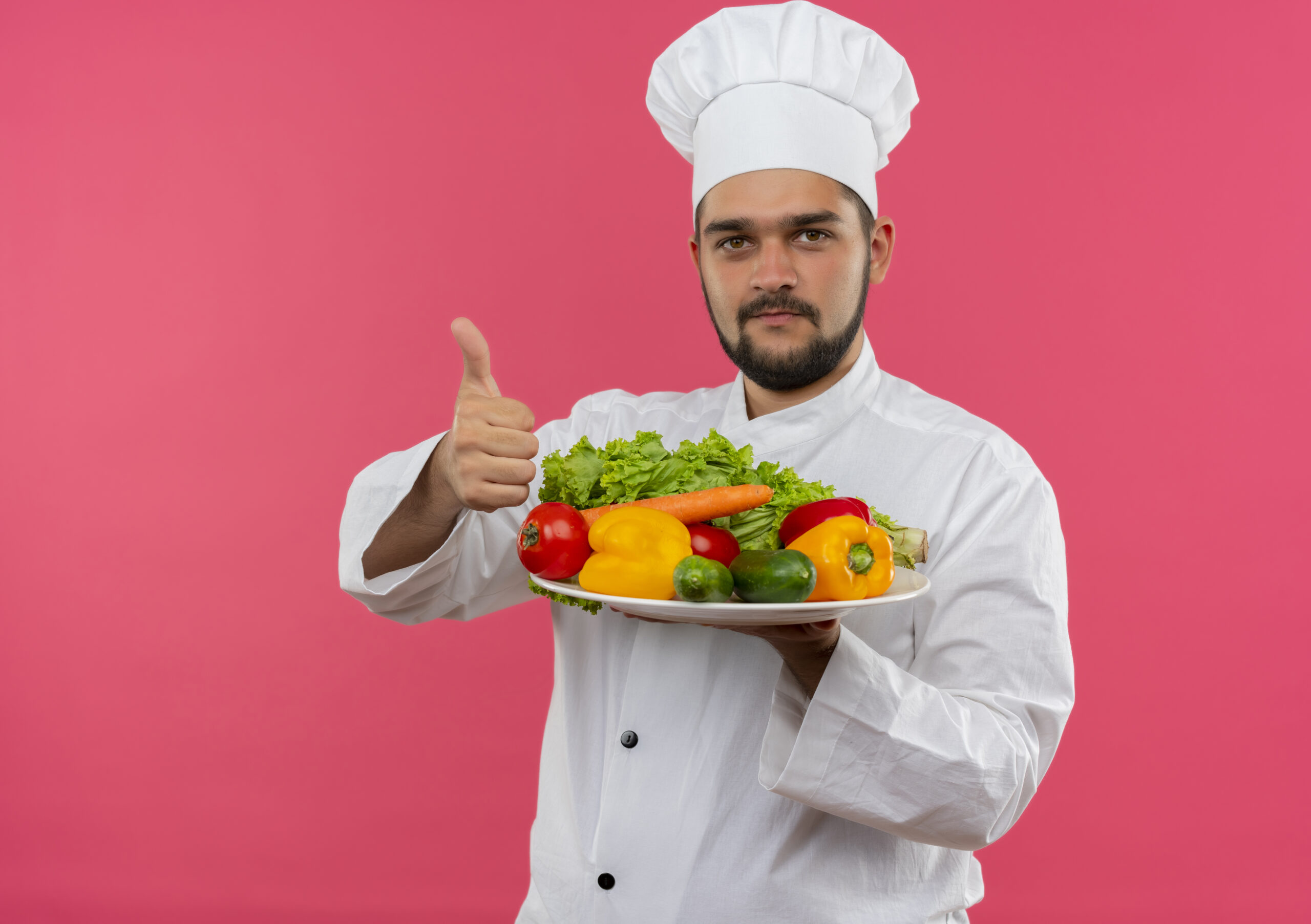 pleased young male cook chef uniform holding plate vegetables showing thumb up isolated pink space scaled